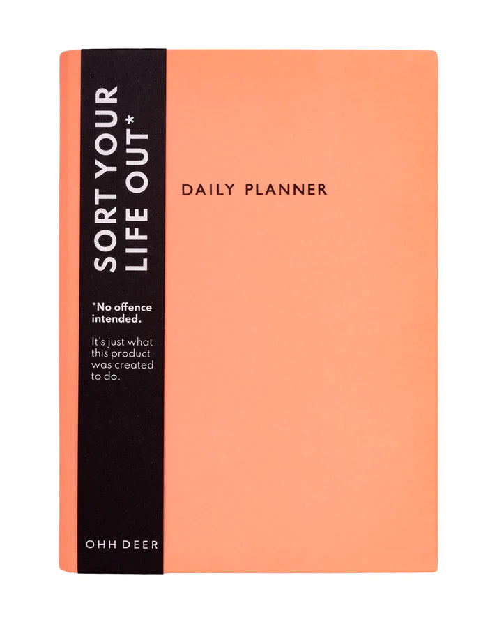 The front cover of a daily planner by Ohh Deer. The cover design is a bright and warm pastel peach or coral colour. There are black embellished words saying in capitals 'DAILY PLANNER'. The image shows a black belly band including details about the product.