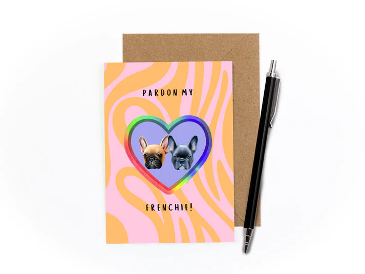 An image of a greetings card with kraft brown envelope tucked into it and a pen sat at an angle to the right. The card is psychedelic in style with swirling light pink and orange in the background. It features a holographic heart sticker in the centre with two cut out heads of French bulldogs - one grey, one brown on a lilac background.