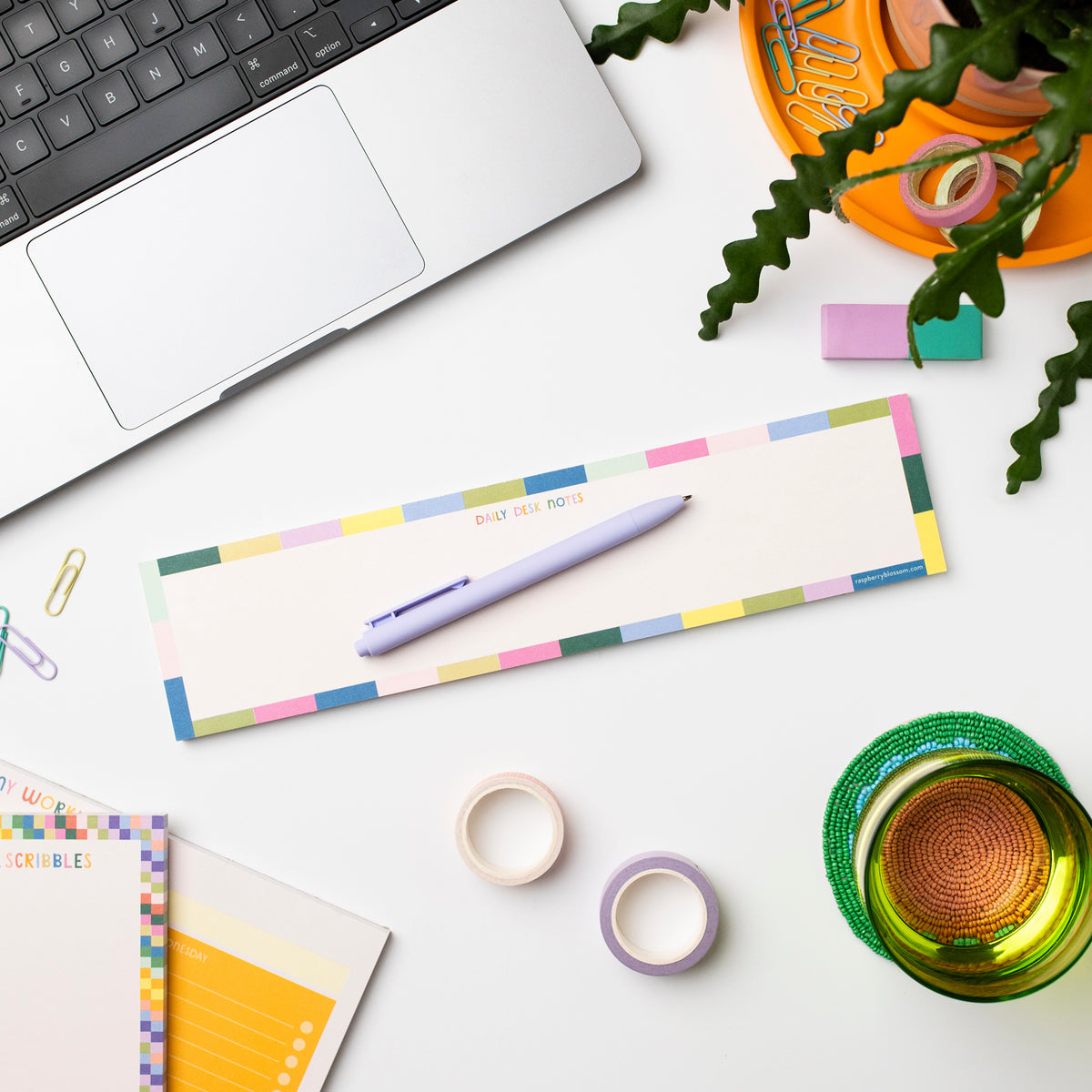 A colourful oblong notepad shown on a desk next to a laptop, other notepads, a glass, a pen, some tape and paperclips plus a pot plant.