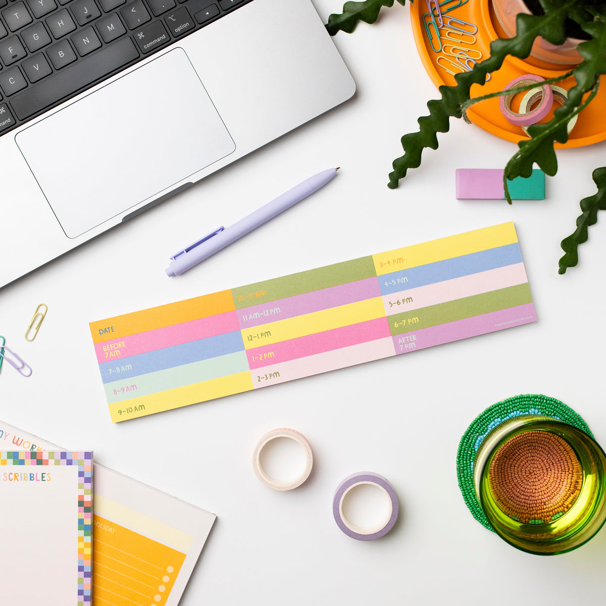 A colourful oblong notepad shown on a desk next to a laptop, other notepads, a glass, a pen, some tape and paperclips plus a pot plant.