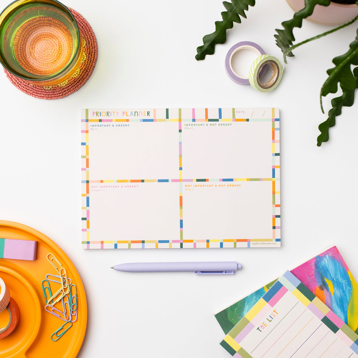 A colourful rectangular notepad shown on a desk next other notepads, a glass, a pen, some tape and paperclips plus a pot plant.