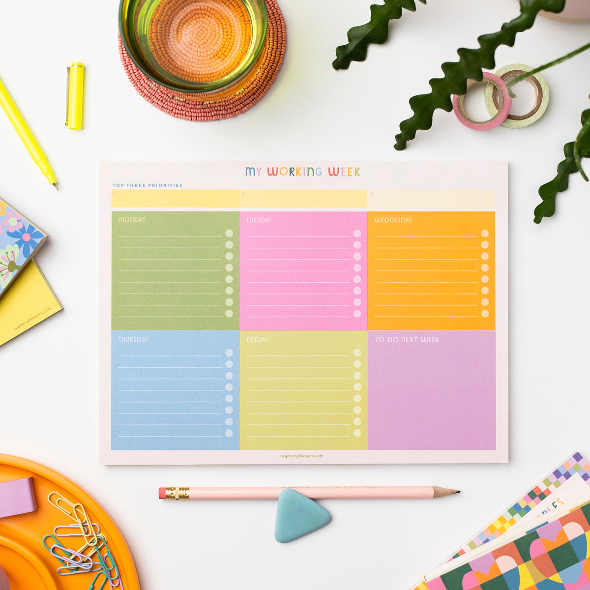 A colourful rectangular notepad shown on a desk next to other notepads, a glass, a pen, some tape and paperclips plus a pot plant.