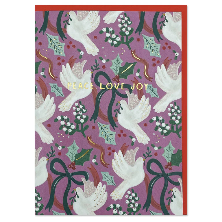 An image of a dusky pink background christmas greetings card. The design is repeat pattern of painted doves, mistletoe, holly and bows. There is gold foiling highlighting areas of the design and words feature in capital gold foiled letters - PEACE, LOVE, JOY across the centre of the card. It has a festive red envelope within the card.