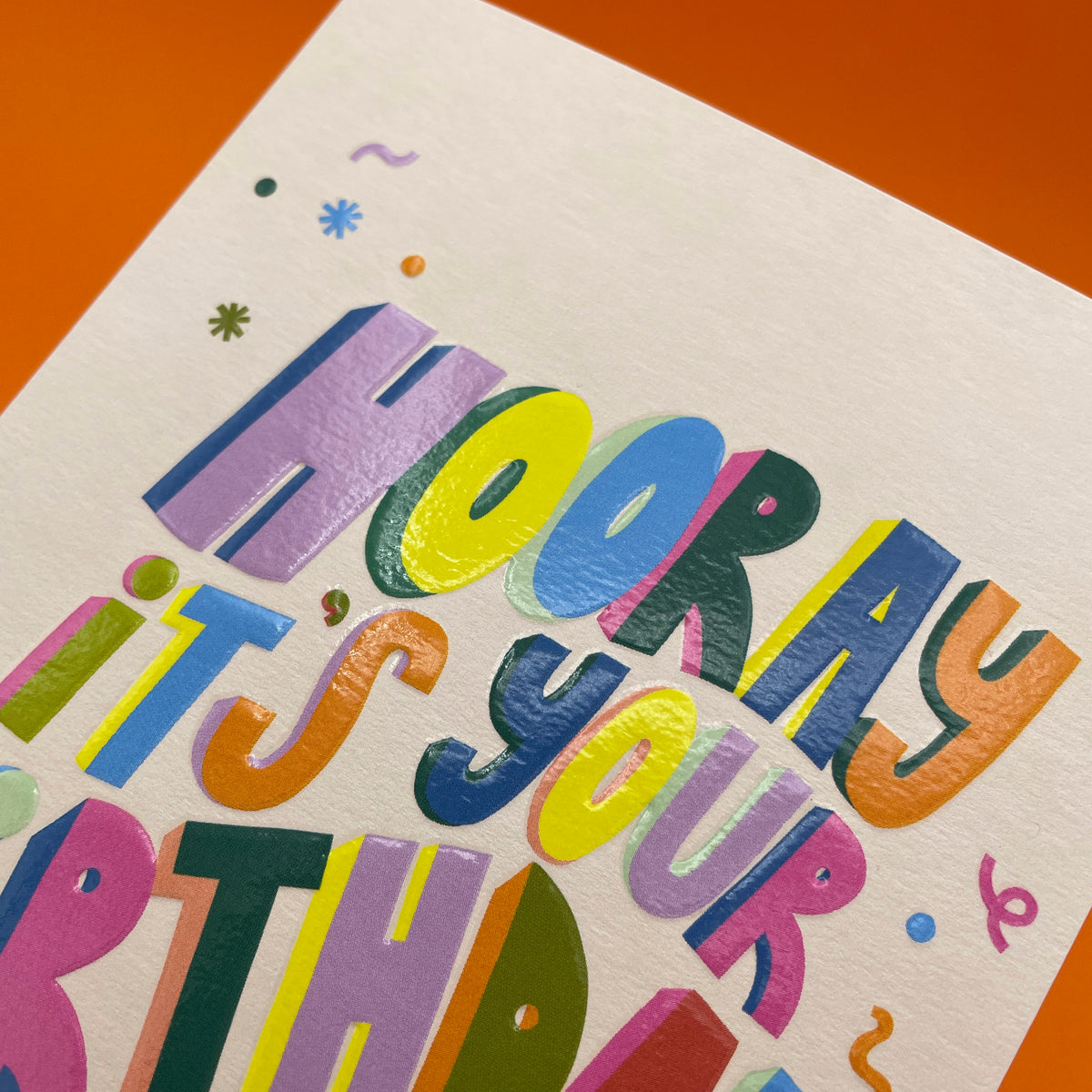 A cream coloured greetings card for a birthday, with a bright orange envelope. It has big wavy colourful words across the front saying in 3D capital letters &#39;hooray it&#39;s your birthday&#39;. There is some confetti flying around the words too.