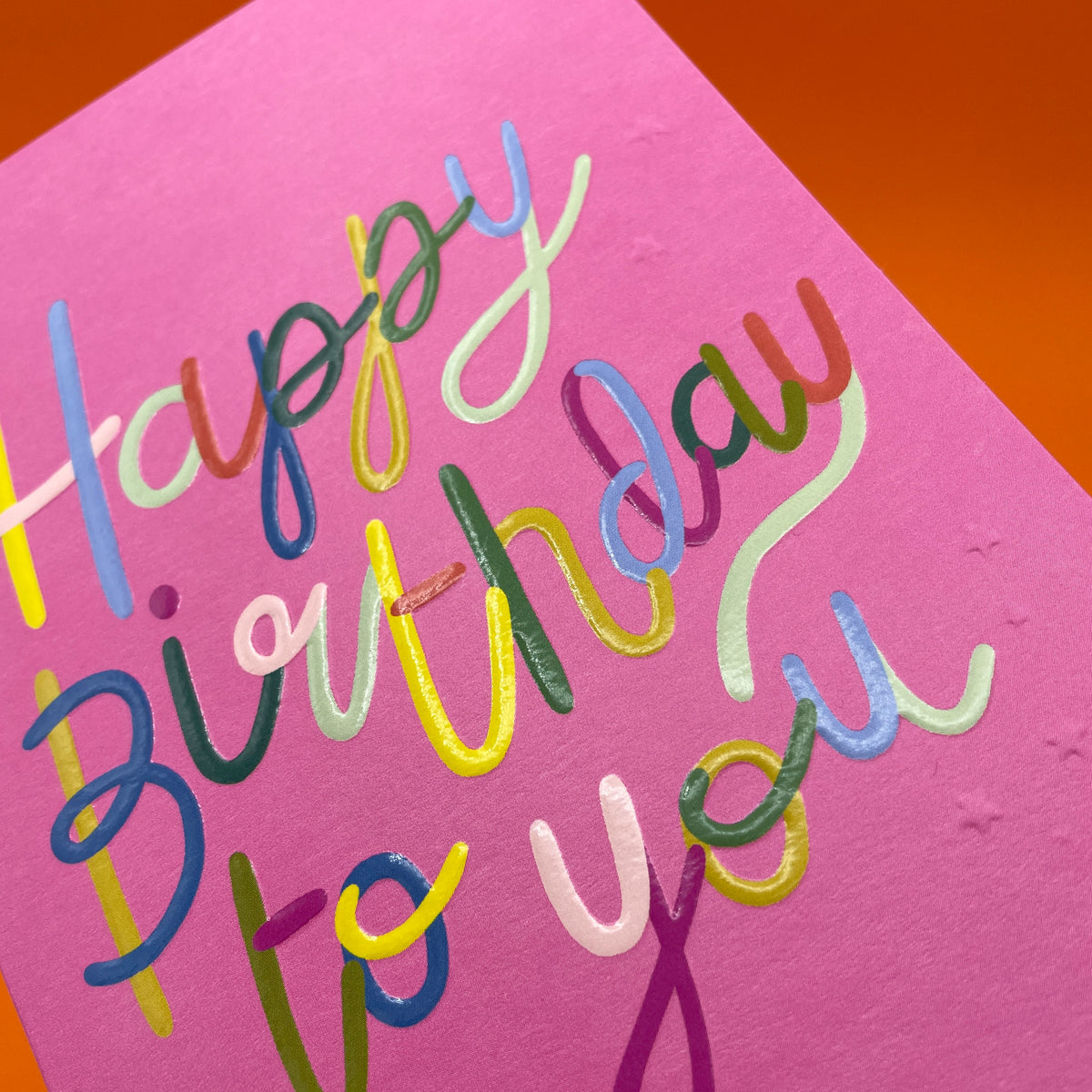 A colourful greetings card to celebrate a birthday where the main colour is hot pink. The wording on the card is &#39;Happy Birthday to you&#39; in big cursive style wrting. Each part of each letter is a different colour.