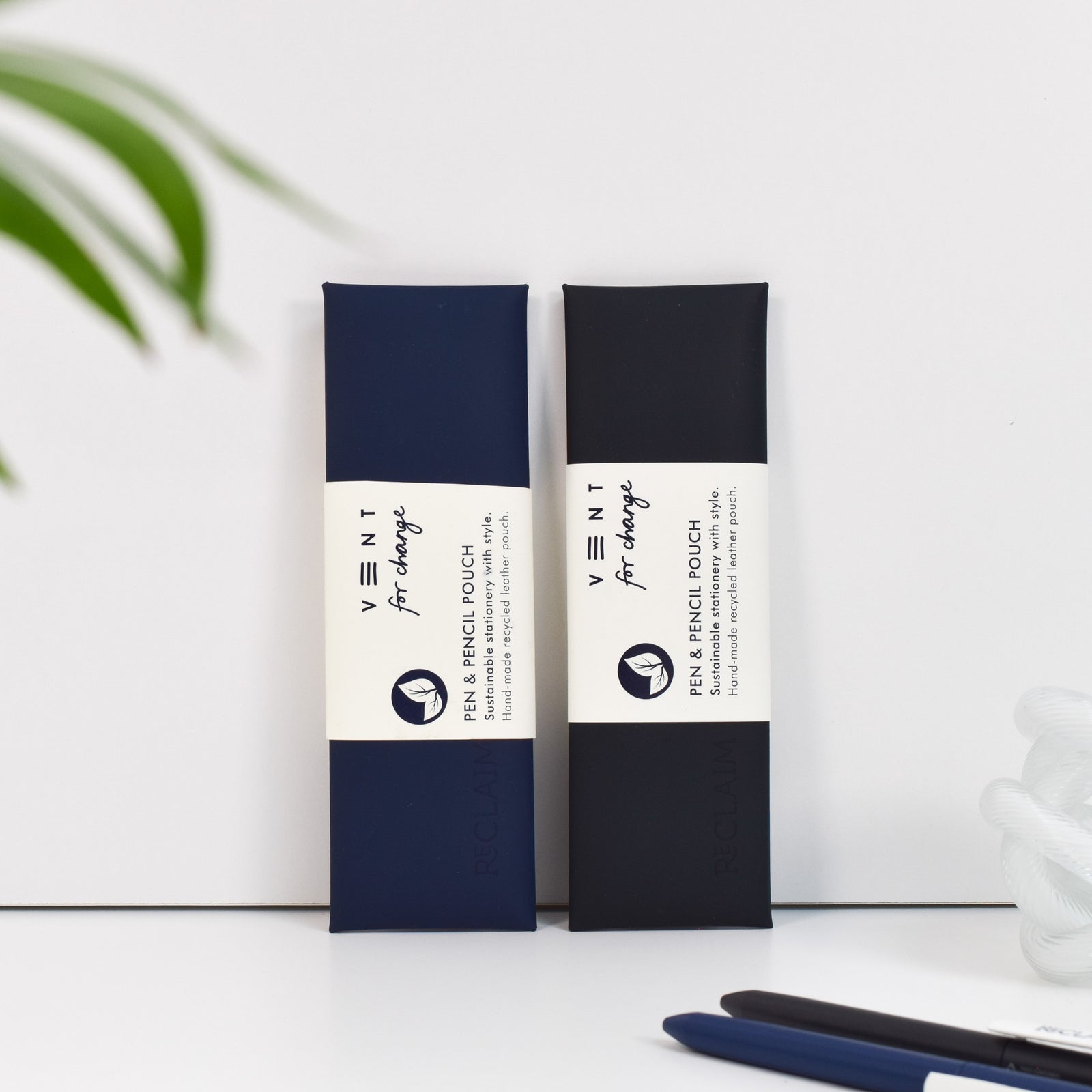 A pair of pen and pencil pouches made of recycled leather with the word 'Reclaim' embossed on them leaning against a wall.