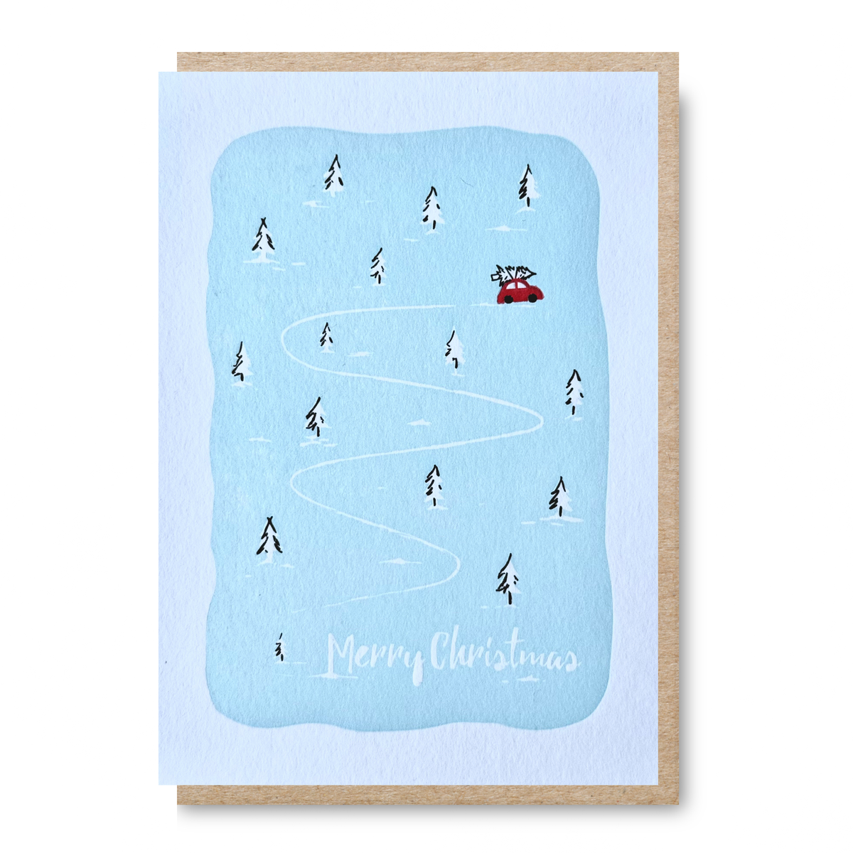 A Christmas greeting card with a light blue snowy winter scene, showing a red car on a winding white path through small black pine trees, with 'Merry Christmas' written at the bottom. The card is shown with a kraft paper envelope behind it on a white background