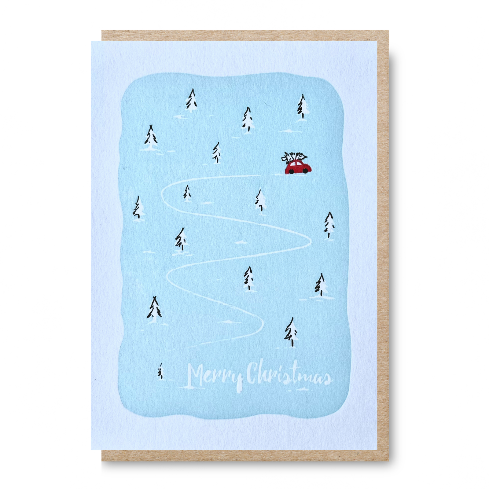 A Christmas greeting card with a light blue snowy winter scene, showing a red car on a winding white path through small black pine trees, with 'Merry Christmas' written at the bottom. The card is shown with a kraft paper envelope behind it on a white background