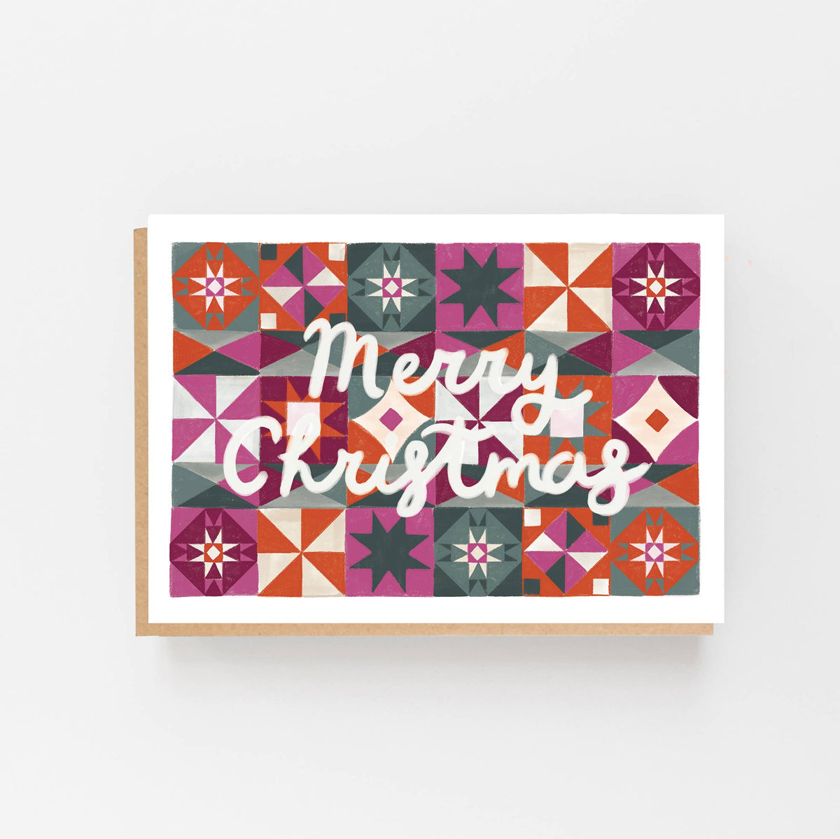 An A6 Christmas greeting card with a colourful geometric quilt pattern background and the words 'Merry Christmas' in white script, shown with a kraft envelope on a light gray background.