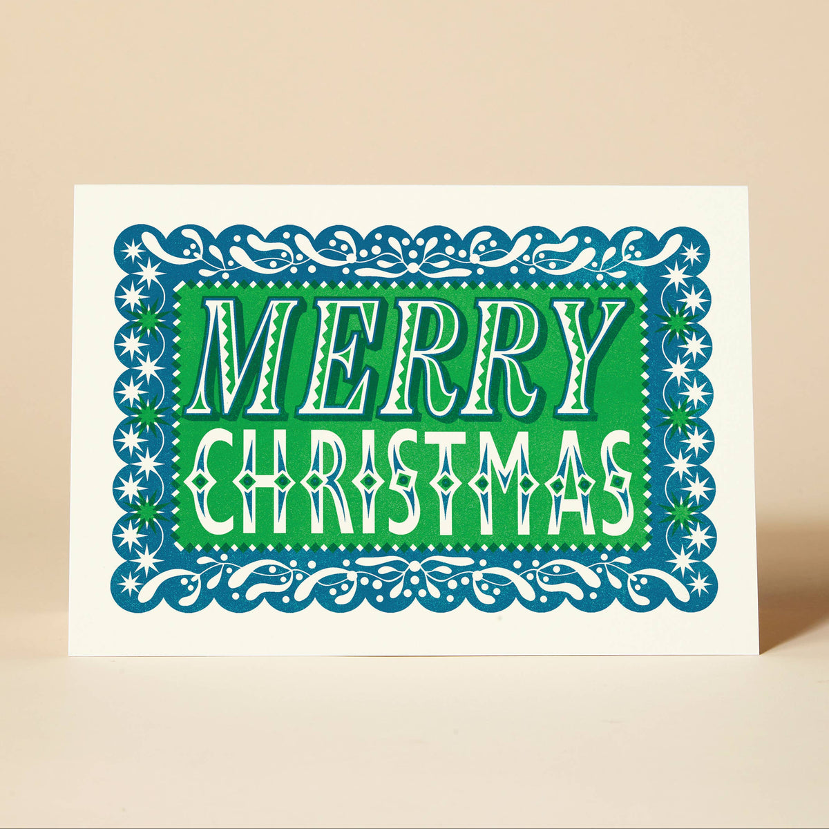 A Christmas greeting card with a white border, featuring a green centre with the words “MERRY CHRISTMAS” in white and a blue ornate decorative border, photographed on a warm beige background.