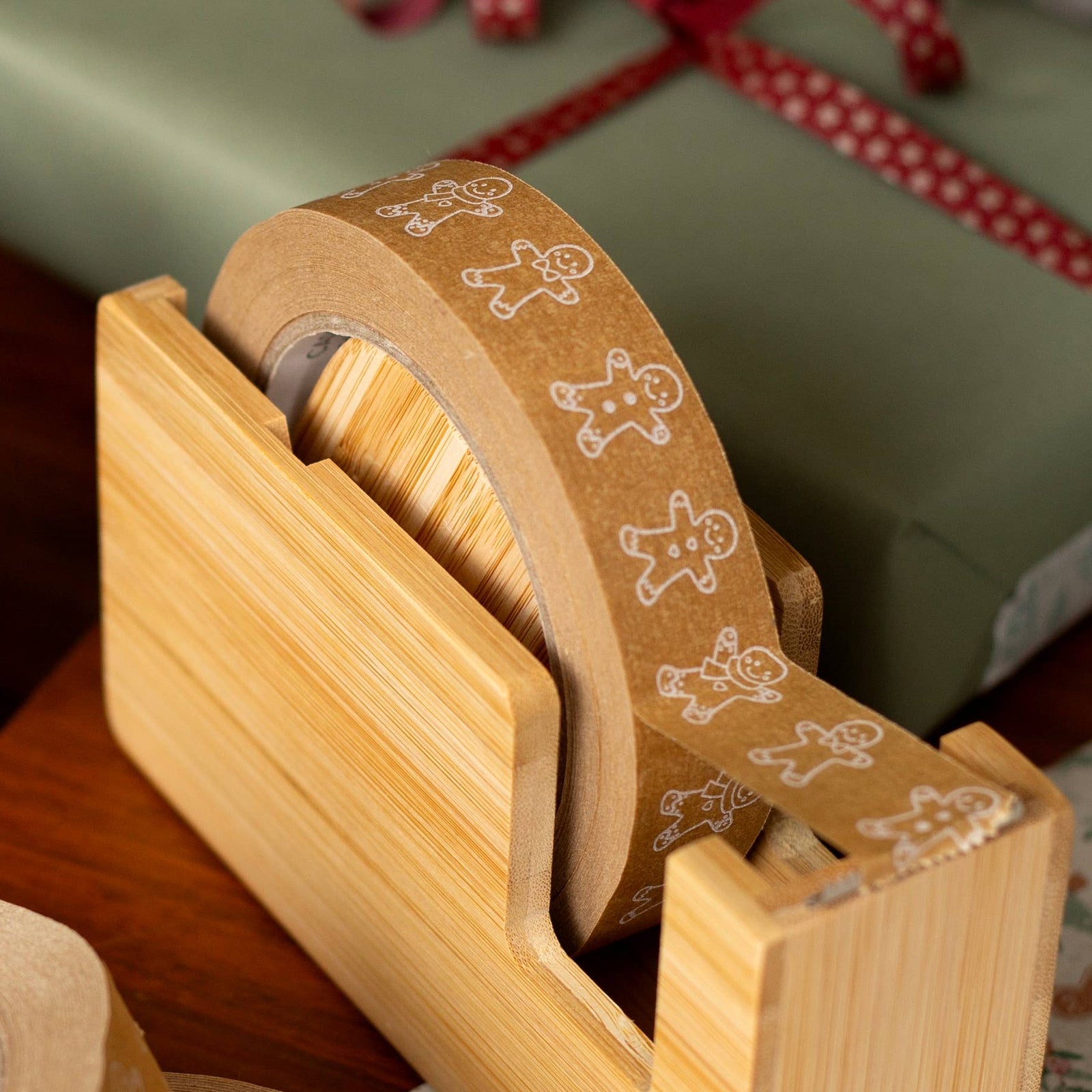 A bamboo gift tape dispenser holding a roll of gingerbread-themed paper packing tape, surrounded by wrapped gift boxes and additional rolls of tape on a wooden table in a holiday setting.