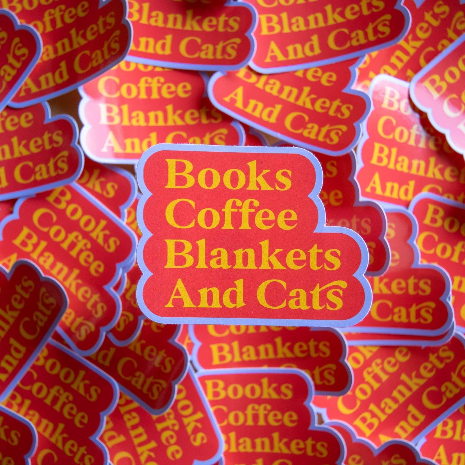 Stack of 'Books Coffee Blankets And Cats' stickers with yellow text on a red background.