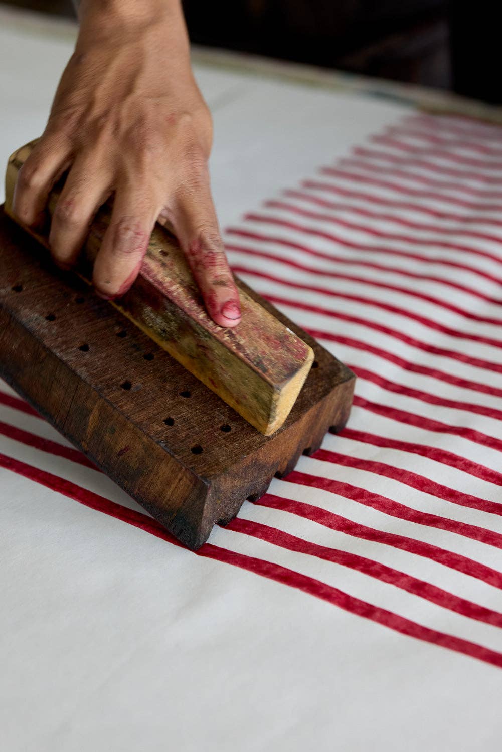 A close-up of a hand using a dark wooden block print press to apply red wax or dye onto white paper, creating a red and white striped pattern.