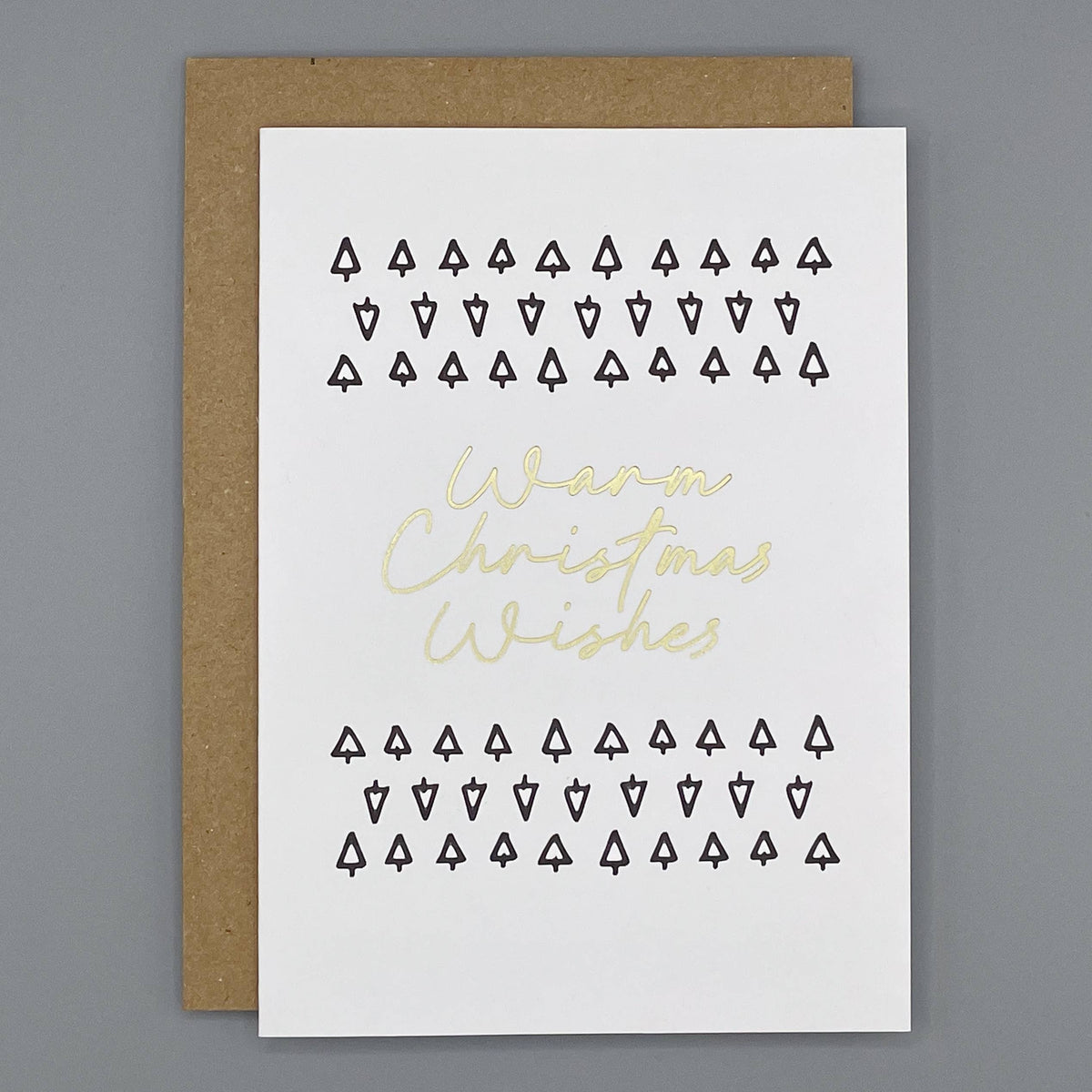 A white Christmas greeting card with black printed rows of small tree icons and gold foil text reading 'Warm Christmas Wishes', shown with a brown kraft envelope on a gray background.