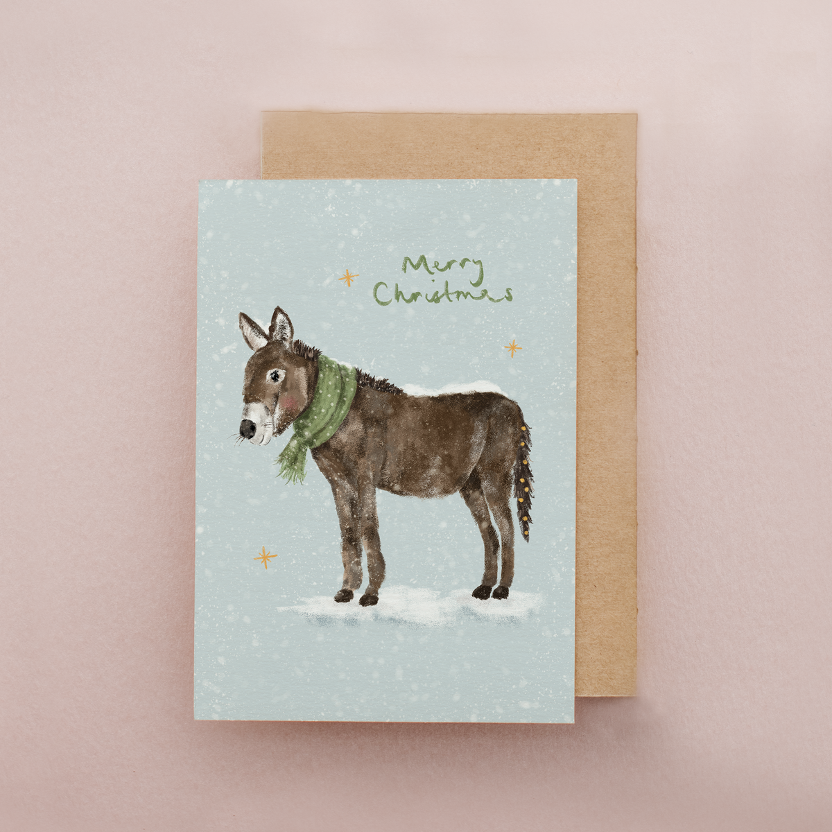 A Christmas greeting card featuring an illustration of a brown donkey wearing a green scarf on a light blue background with the words 'Merry Christmas' and small gold stars, shown with a kraft envelope on a dusty pink background.