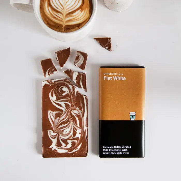 A white table top with a white coffee mug featuring latte art. at the top of the image. In the middle are two chocolate bars - one with the wrapper on and one off. The naked chocolate bar is dark brown in colour with white swirls decorating the surface.