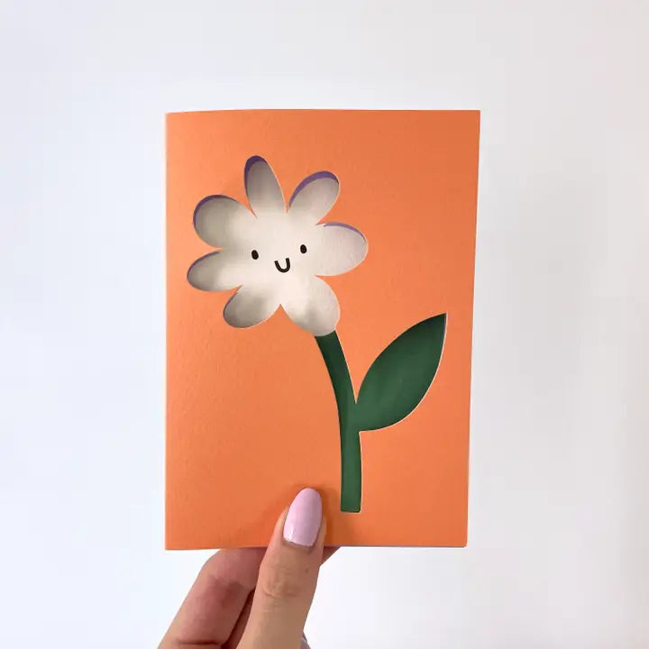 A greetings card with an orange background and the image of a flower where the head of the flower is cut out of the card and the flower's smiling face lies behind.