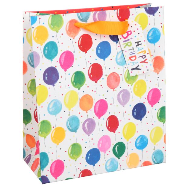 A gift bag with a white background covered in a rainbow of coloured balloons with strings dangling. The white background also has some multicoloured spots embellishing it. There is an orange ribbon handle with a gift tag attached with the words Happy Birthday in a haphazard type.