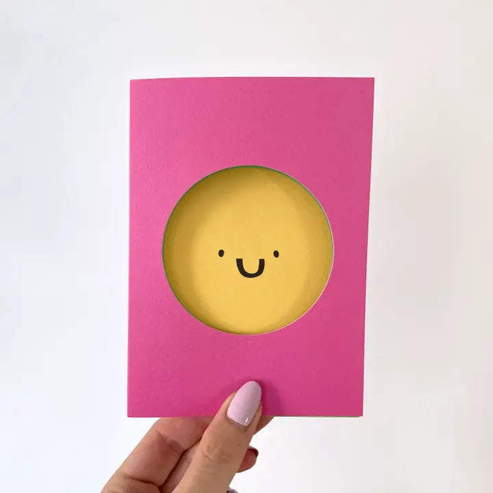 A greetings card with a pink background and the image of a yellow happy face is cut out of the card and the smiling face lies behind.