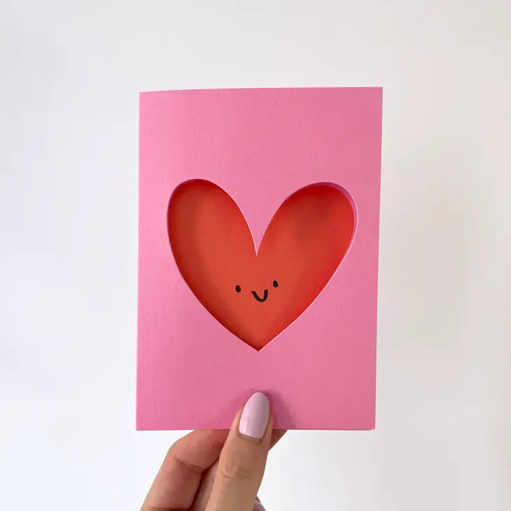 A greetings card with a pink background and the image of a heart is cut out of the card and the heart's smiling face lies behind.