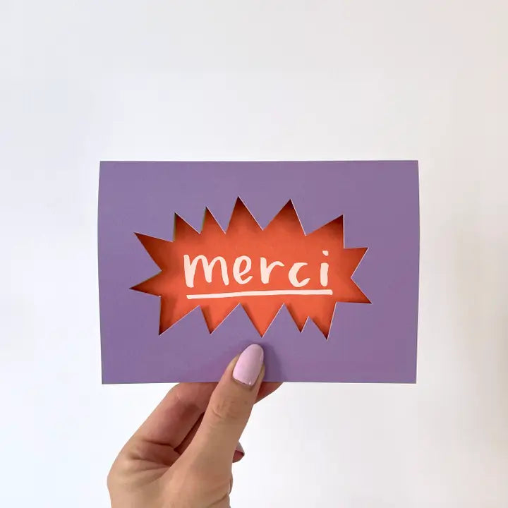 A greetings card with a purple background and an explosive cut out reveals the word 'merci' lies behind.