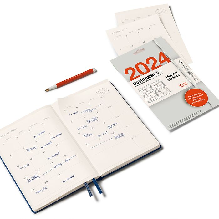 A lifestyle image of an open notebook and a set of 2024 planner stickers open to the right hand side of it. They are laid out on the diagonal. The notebook has the stickers stuck onto the pages and there are a few loose leaf stickers in the shot plus an orange pen.
