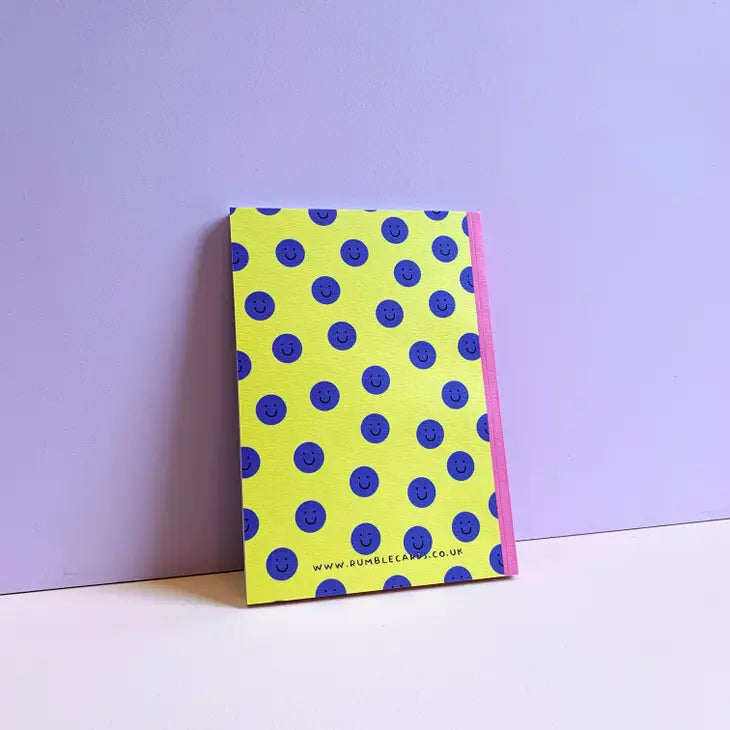 The back of a notebook which is yellow in colour and covered in navy blue circles with happy faces in them. The spine is pink,