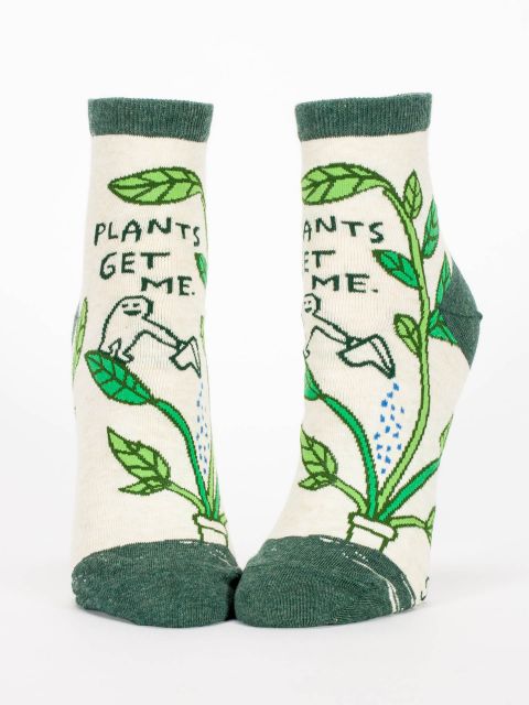 A pair of cream socks with dark green toes, heels and top edge. The image on the socks is a big green potted plant being watered by a being and the words &#39;Plants Get Me&#39;.