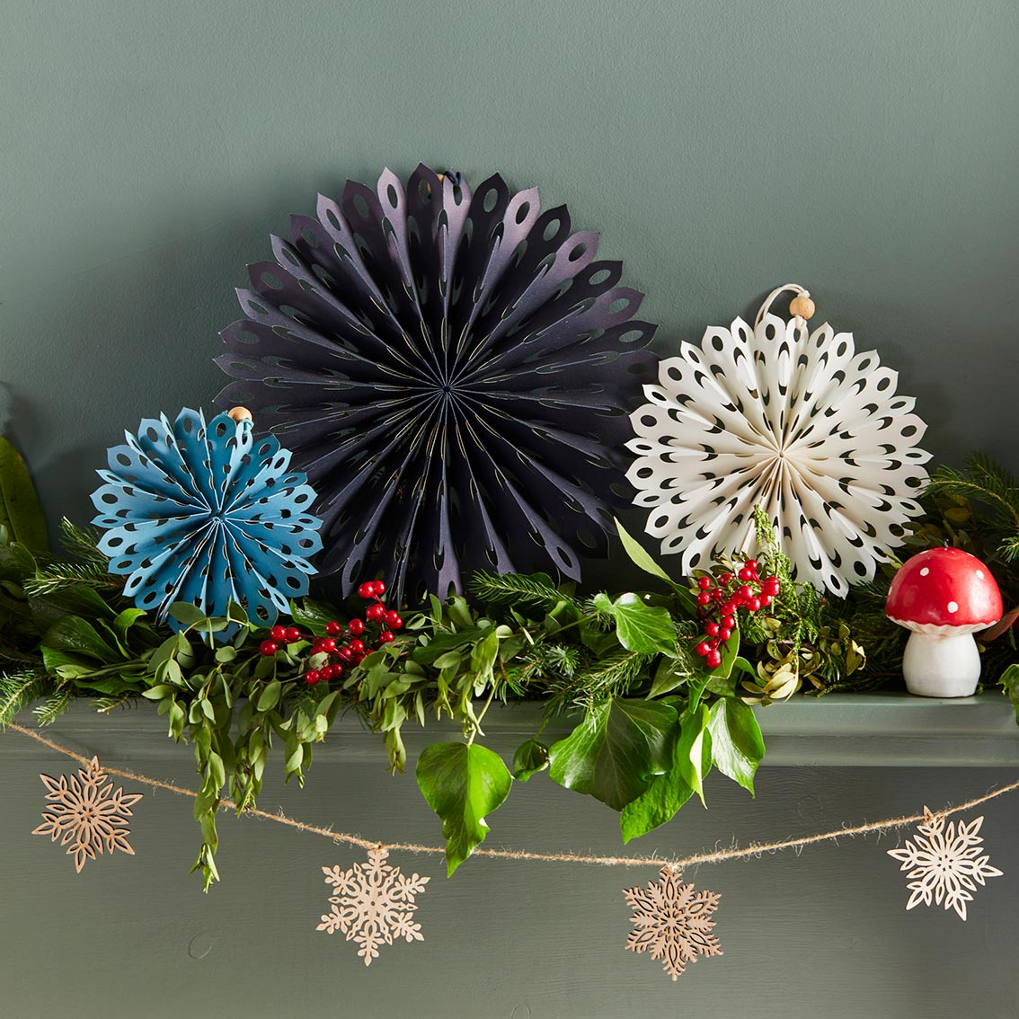 An image of a mantlepiece decorated for Christmas with 3 paper honeycomb fans, some wooden snowflake bunting, a toadstool candle and a foliage garland.