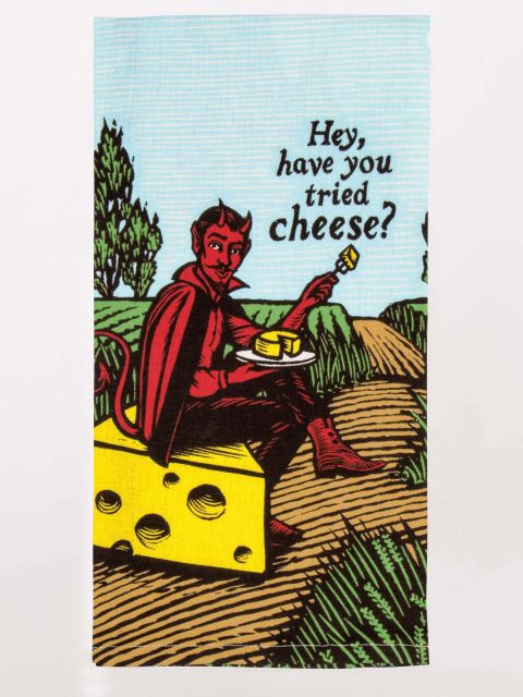 A folded tea towel showing the design of a red devil in a field sitting on a wedge of cheese, offering you a wedge of cheese by asking 'hey, have you tried cheese?'