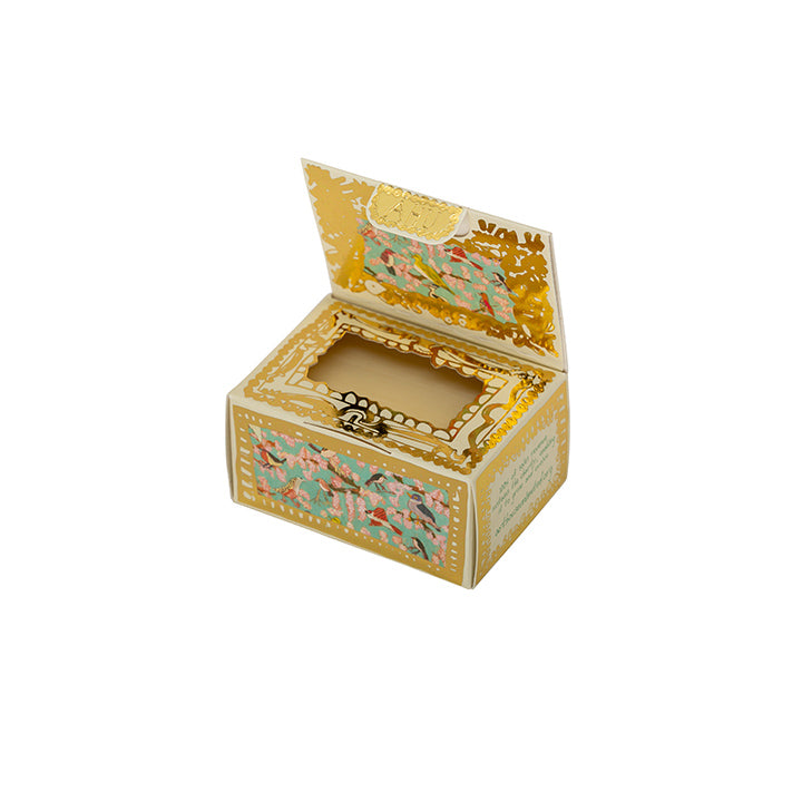 A cuboid shape box with guilded gold edging and a bright red/pink label stating the company Arthouse Unlimited. There is a background illustration of colourful birds. The box lid is open revealing a soap inside.