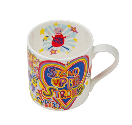 A white china mug with a rainbow coloured artwork of a large heart and the words stand up be strong on the outside and on the inside, a smaller image of a white person with the words smile inside on their top.