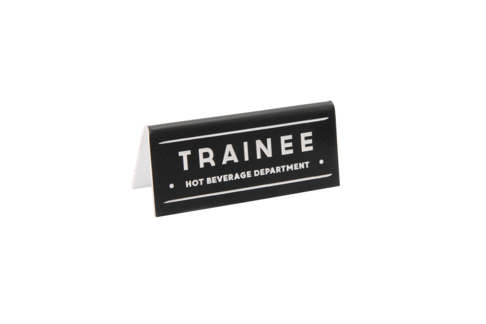 A small black acrylic sign that has in big white capital letters on it &#39;TRAINEE HOT BEVERAGE DEPARTMENT&#39;.