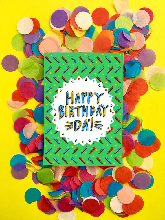 A greetings card with a grass green background detailed with red and blue markings and a white circular cut out in the middle with the capitalised blue lettering HAPPY BIRTHDAY DA&#39;