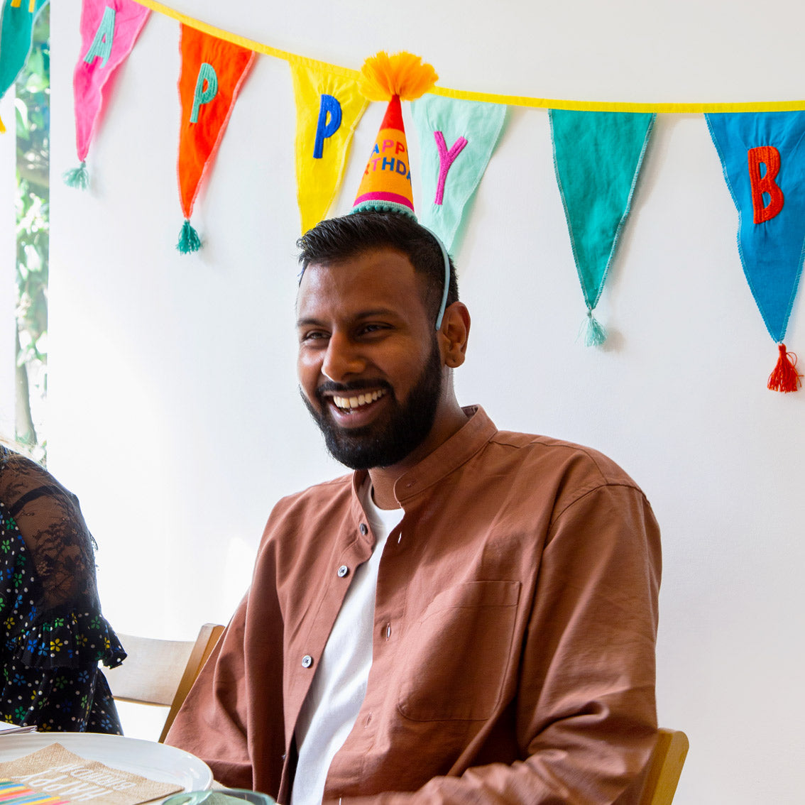 A person of colour at a party wearing a cone shape birthday headband with rainbow fabric bunting in the background.