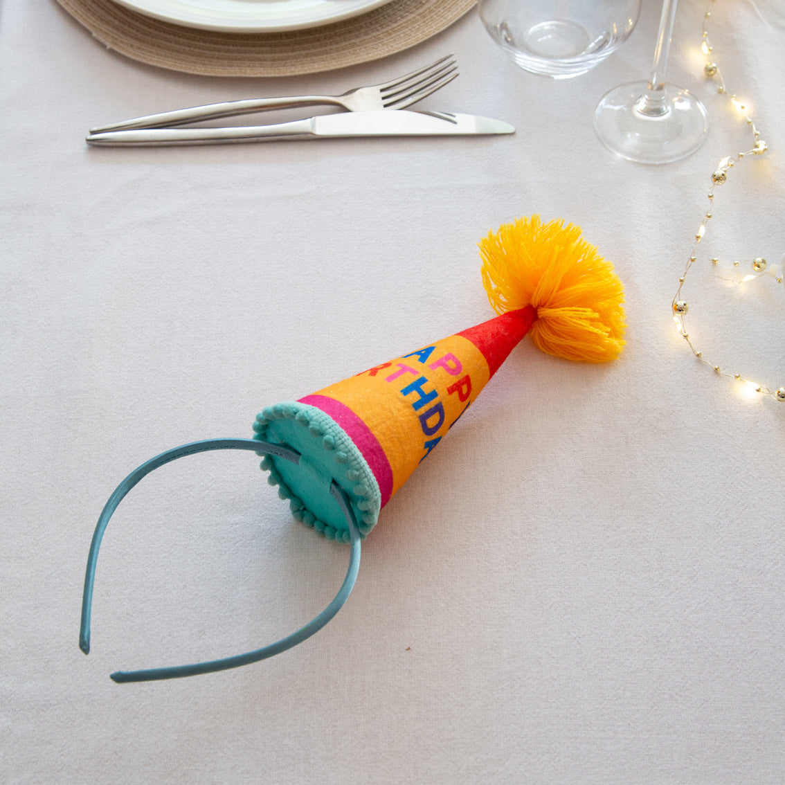 Blue headband with traditional cone party hat on top with pompom on top, featuring the words happy birthday, lying on a party table.