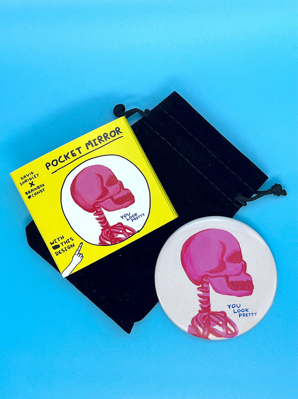 A white round pocket mirror with drawing of a pink skeleton from the shoulders and navy handwriting saying You Look Pretty by artist DAvid Shrigley. There is a black velvet drawstring pouch that is comes in and bright yellow packaging to match.