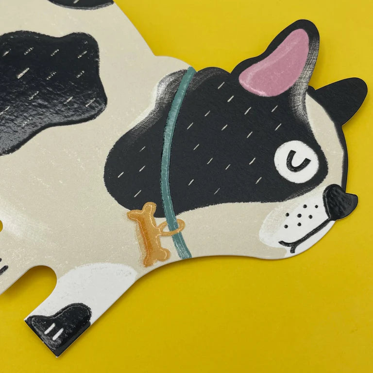 A card cut out in the shape of a black and beige patchy French Bulldog with it&#39;s eyes closed. The dog is standing side facing and has a bone shape attached to it&#39;s green colour. A yellow envelope is behind the dog.