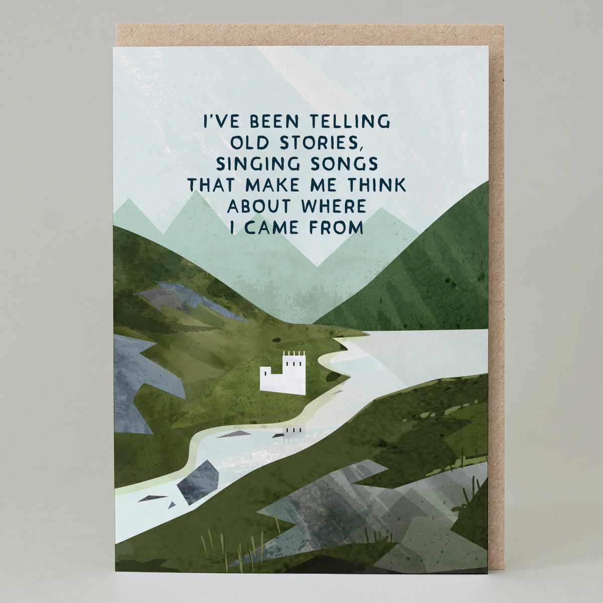 A greetings card featuring a green landscape similar to Scotland, with a white castle on the edge of a loch and a mountain range in the distance. It features the words in dark green capitals from the famous song - I&#39;ve been telling old stories, singing songs that make me think about where I come from.