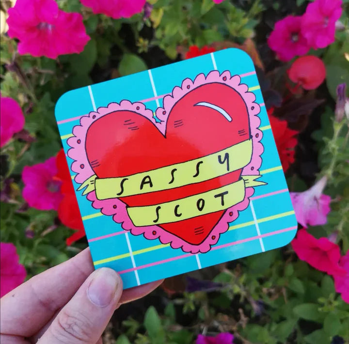 A sky blue coaster with a light tartan design in the background, featuring a big red filly heart with a banner saying SaSSY SCOT in yellow across the middle.