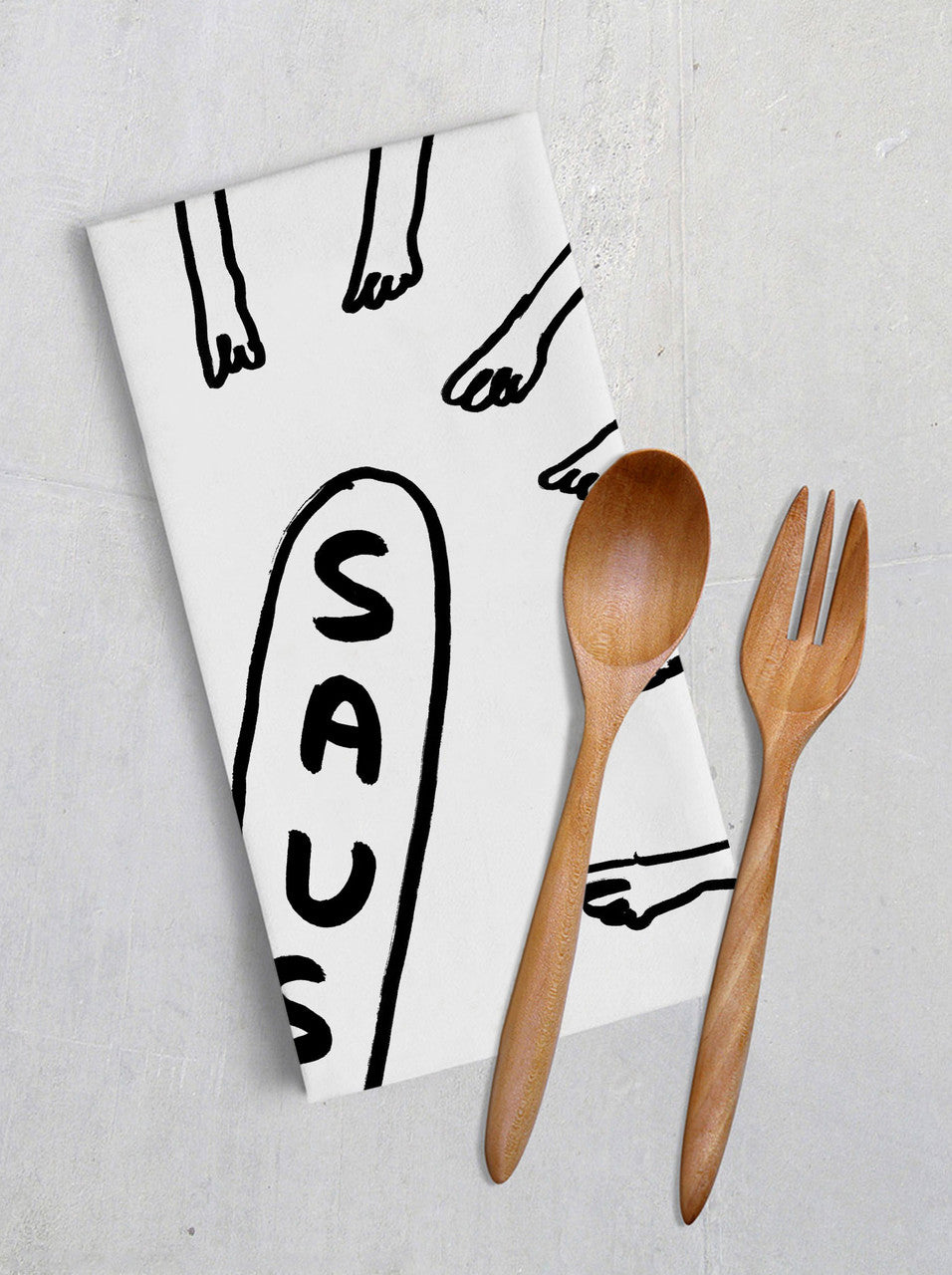 A white tea towel with a black outlined illustration of many hands pointing to a sausage. The sausage is the outline shape with the word SAUSAGE in block capitals. This is a lifestyle image with the tea towel folded alongside wooden kitchen implements.
