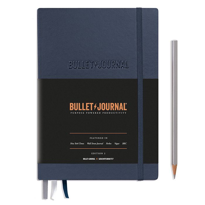 Navy coloured notebook with band keeping it closed. It features a paper band describing it&#39;s a bullet journal layout inside. A pencil sits alongside it.