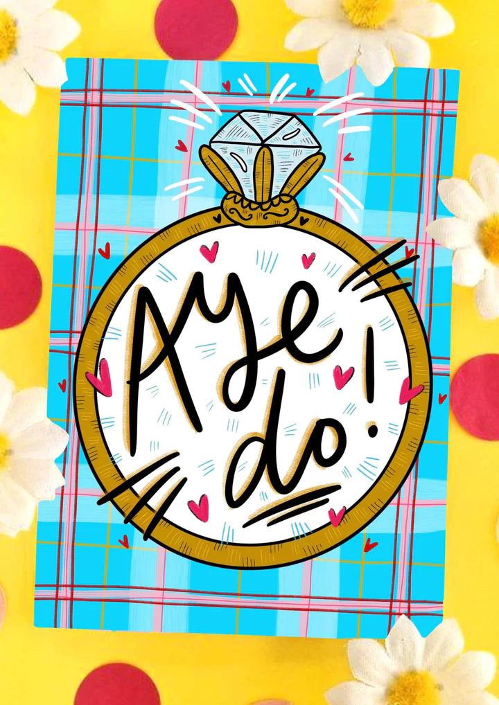 A greetings card with blue, pink and red tartan background, featuring a large diamond ring and the words AYE DO in the middle.